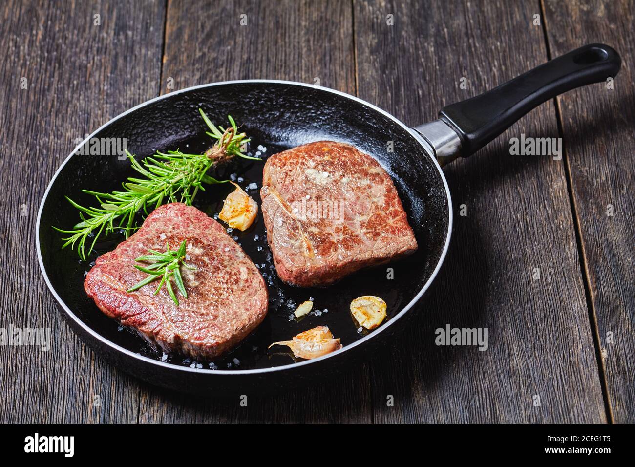 steaks de bœuf frits au bouquet de romarin, ail et sel sur une poêle sur une table en bois sombre, vue horizontale d'en haut Banque D'Images