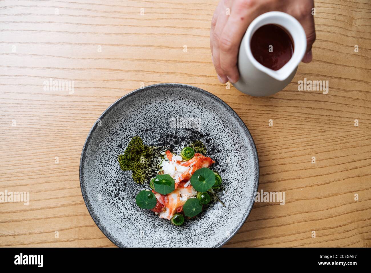 Vue rognée de la main de l'homme en versant de la sauce épaisse rouge pichet dans un bol avec salade hachée, légumes et poisson servi sur une table en bois Banque D'Images