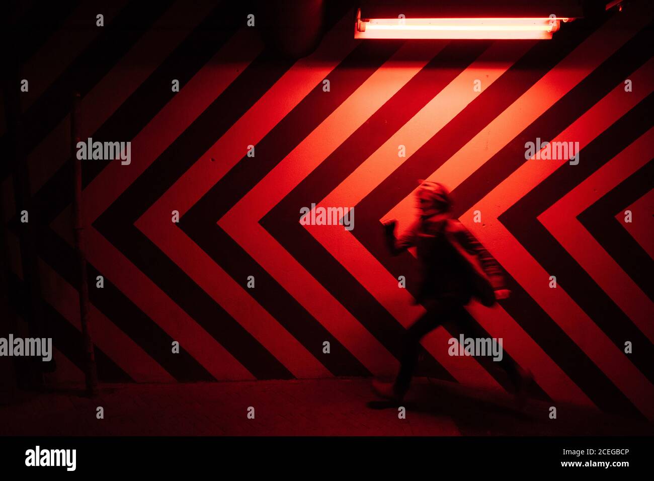 Vue latérale du mouvement représentation floue d'un homme qui marche vers le bas dans le tunnel dans la direction opposée aux grands rouges et noirs flèches sur le mur éclairées par des lampes rouges Banque D'Images