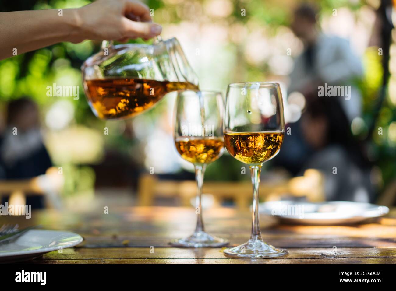 Rognez le vin blanc versé à la main dans des verres à l'aide d'un pichet en verre avec des tiges hautes debout sur une table en bois?rue?café sur le soleil flou arrière-plan Banque D'Images