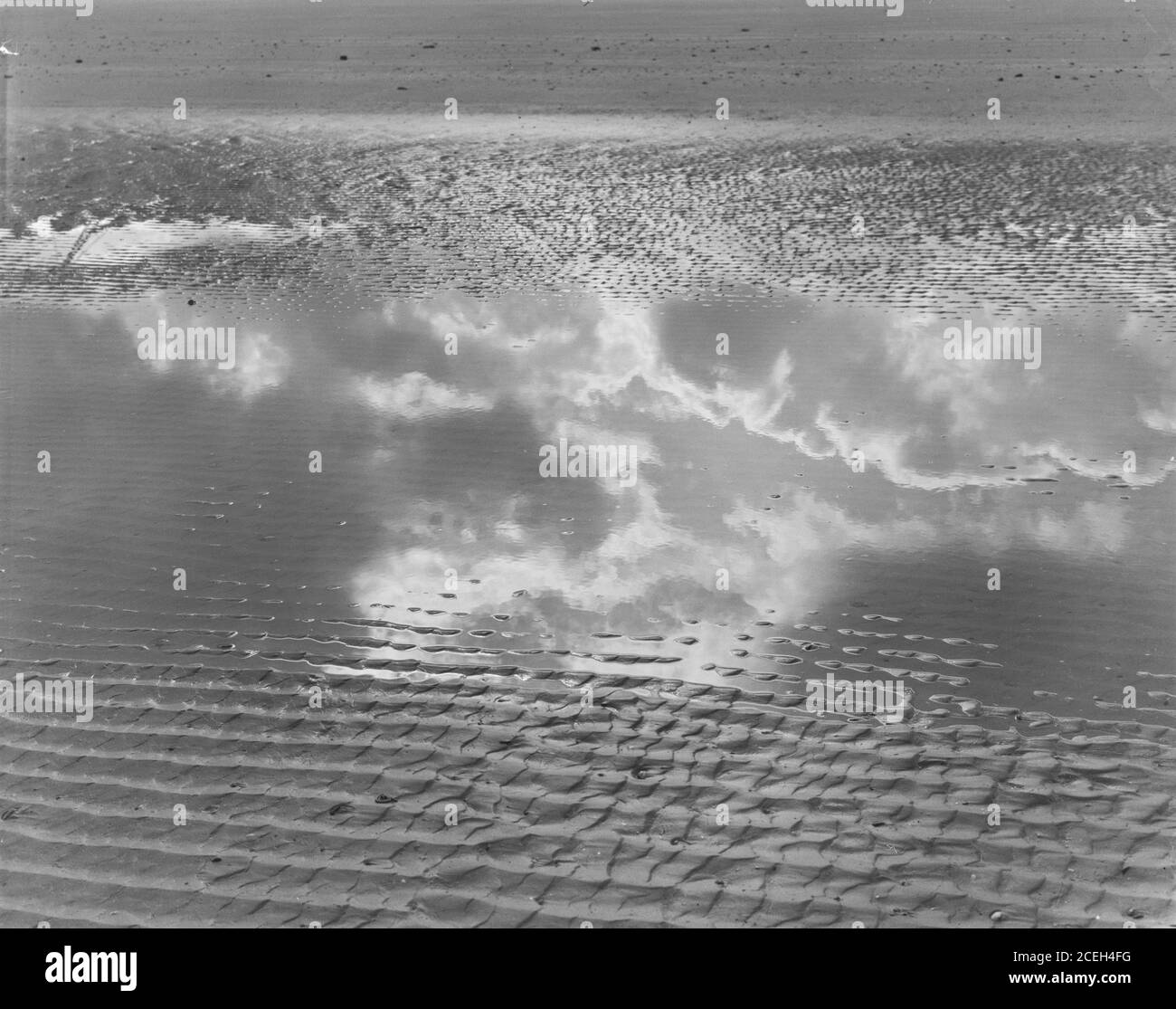 Photo en noir et blanc d'eau peu profonde qui coule sur une plage de sable texturé reflétant les nuages dans le ciel, Belgique. Banque D'Images