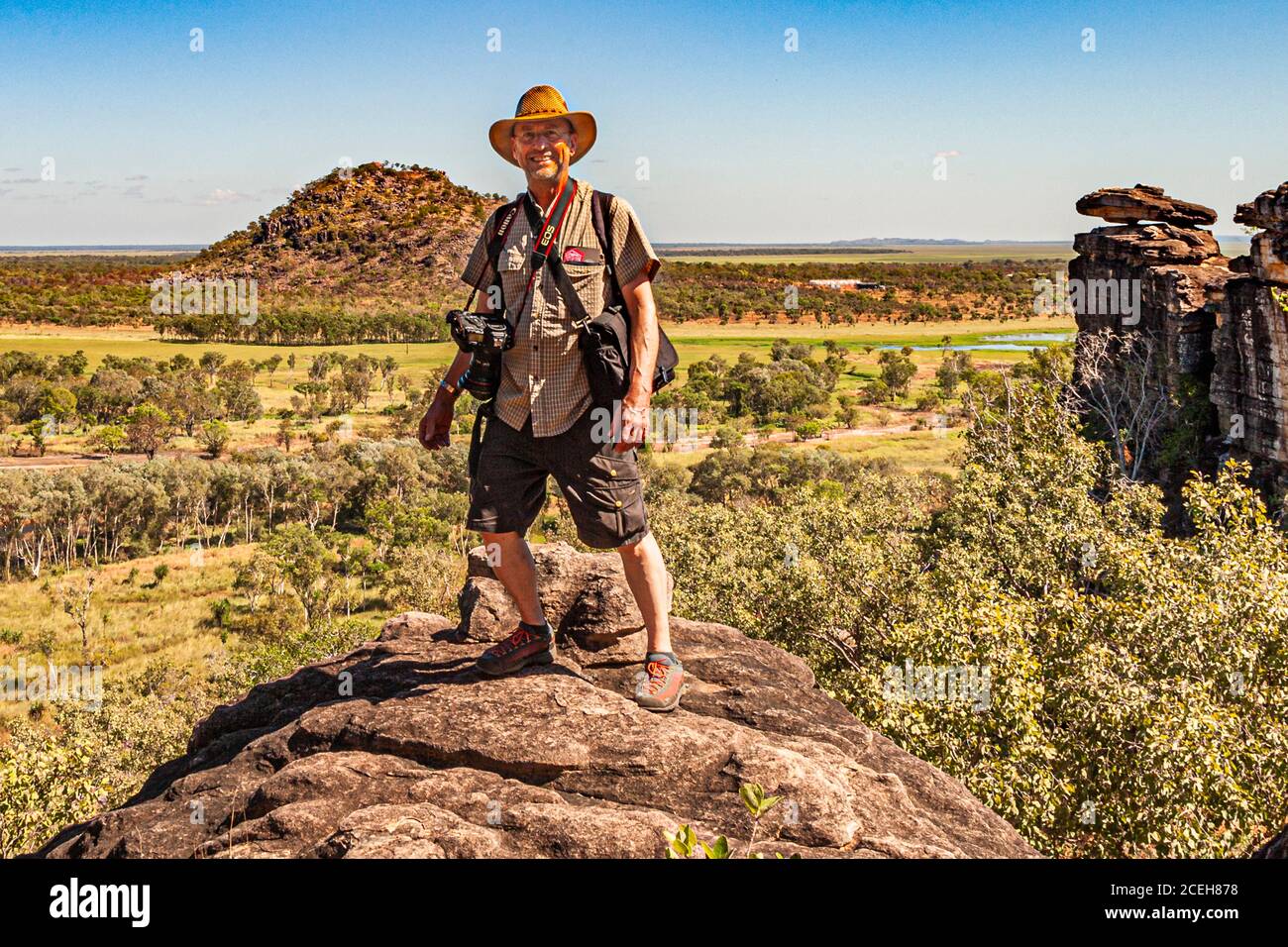 Le photographe Georg Berg se tient avec son sac à dos et son appareil photo sur un rocher près de Point Stuart, dans le Territoire du Nord, en Australie, avec la plaine et une colline de grès solitaire s'étendant derrière lui. Banque D'Images