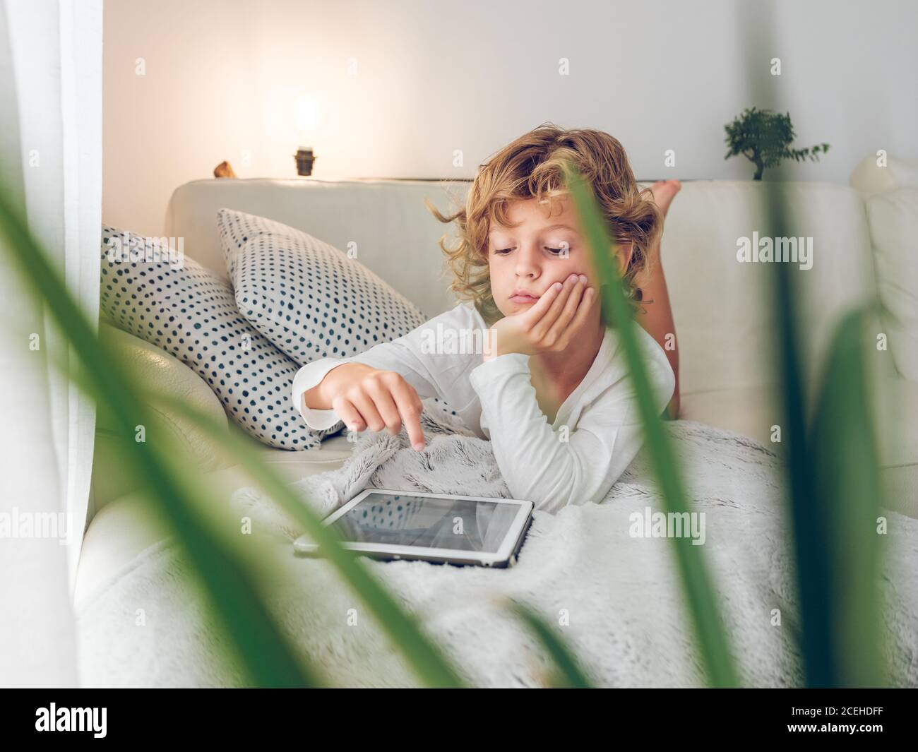 Garçon ennuyé avec des cheveux ondulés pendu tête sur la main et un écran tablette moderne tout en étant allongé sur un canapé confortable Banque D'Images