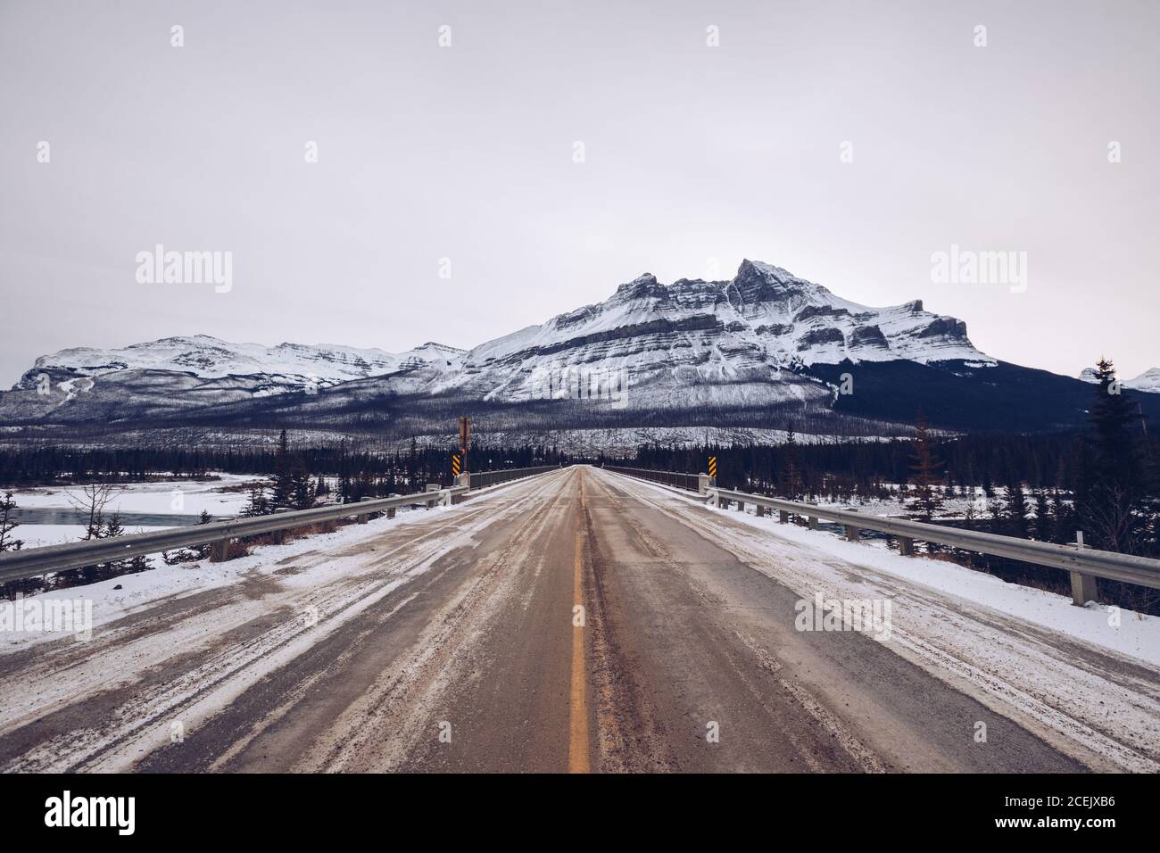 Pont clôturé avec des panneaux qui passent à travers les bois d'hiver épais et rivière gelée sur fond de montagne enneigée avec ciel gris nuageux Banque D'Images