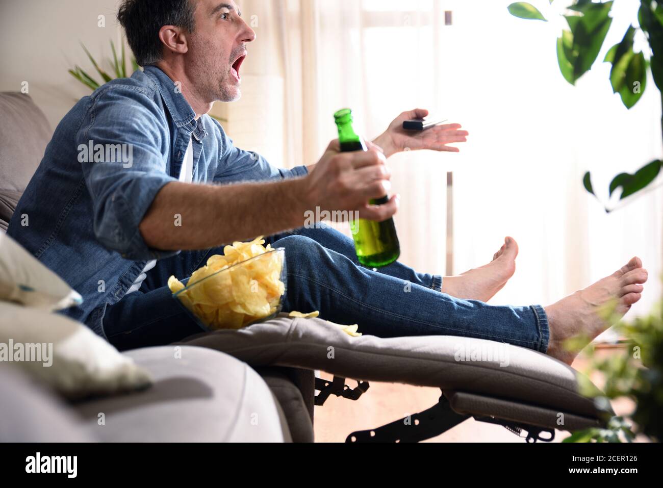 Homme en colère regardant des sports à la télévision assis confortablement sur un canapé avec un en-cas avec de la bière et une télécommande dans ses mains. Banque D'Images