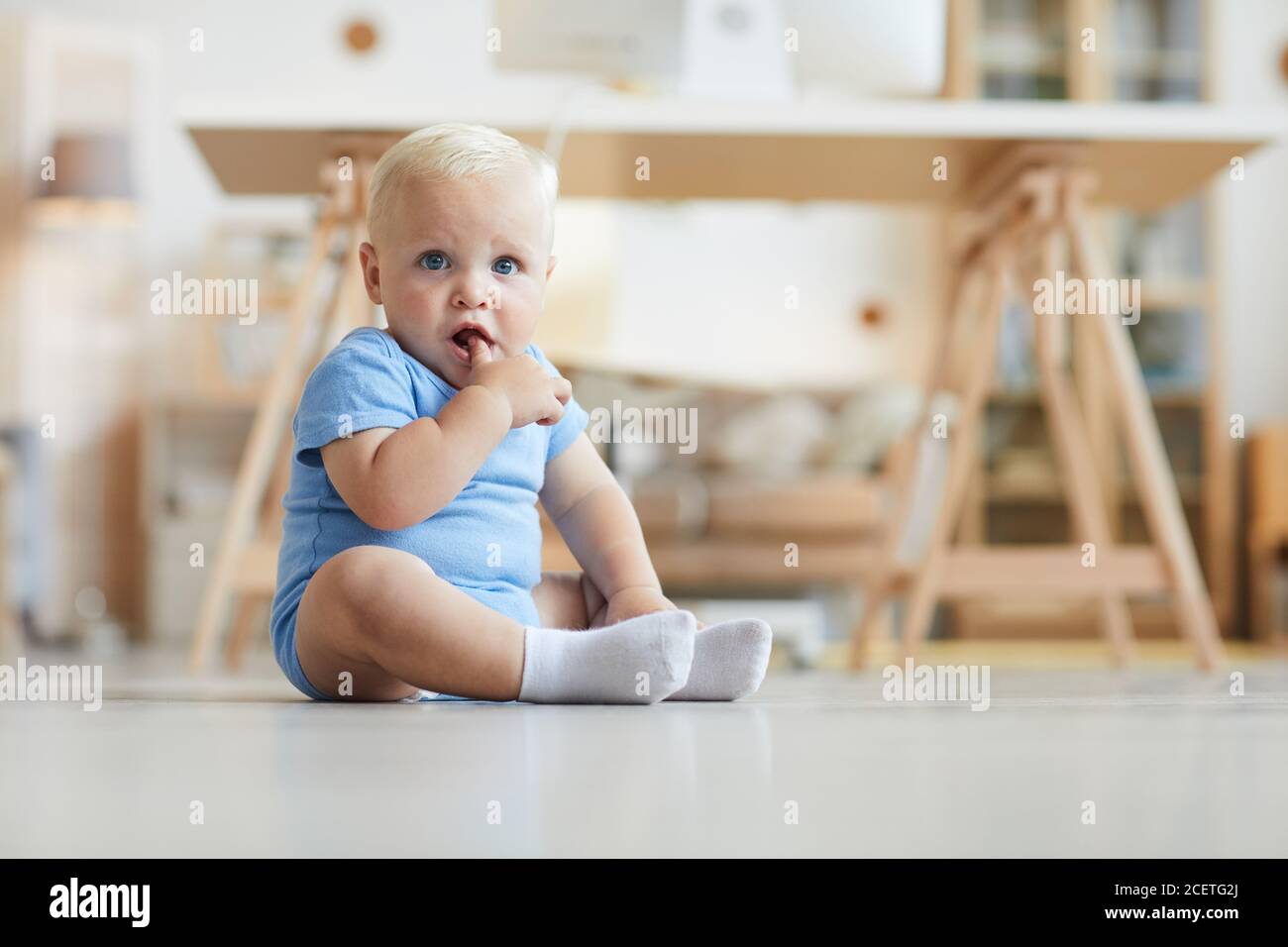 Bébé mignon avec les cheveux blonds et les yeux bleu vif assis sur le sol avec le doigt dans la bouche regardant la caméra Banque D'Images