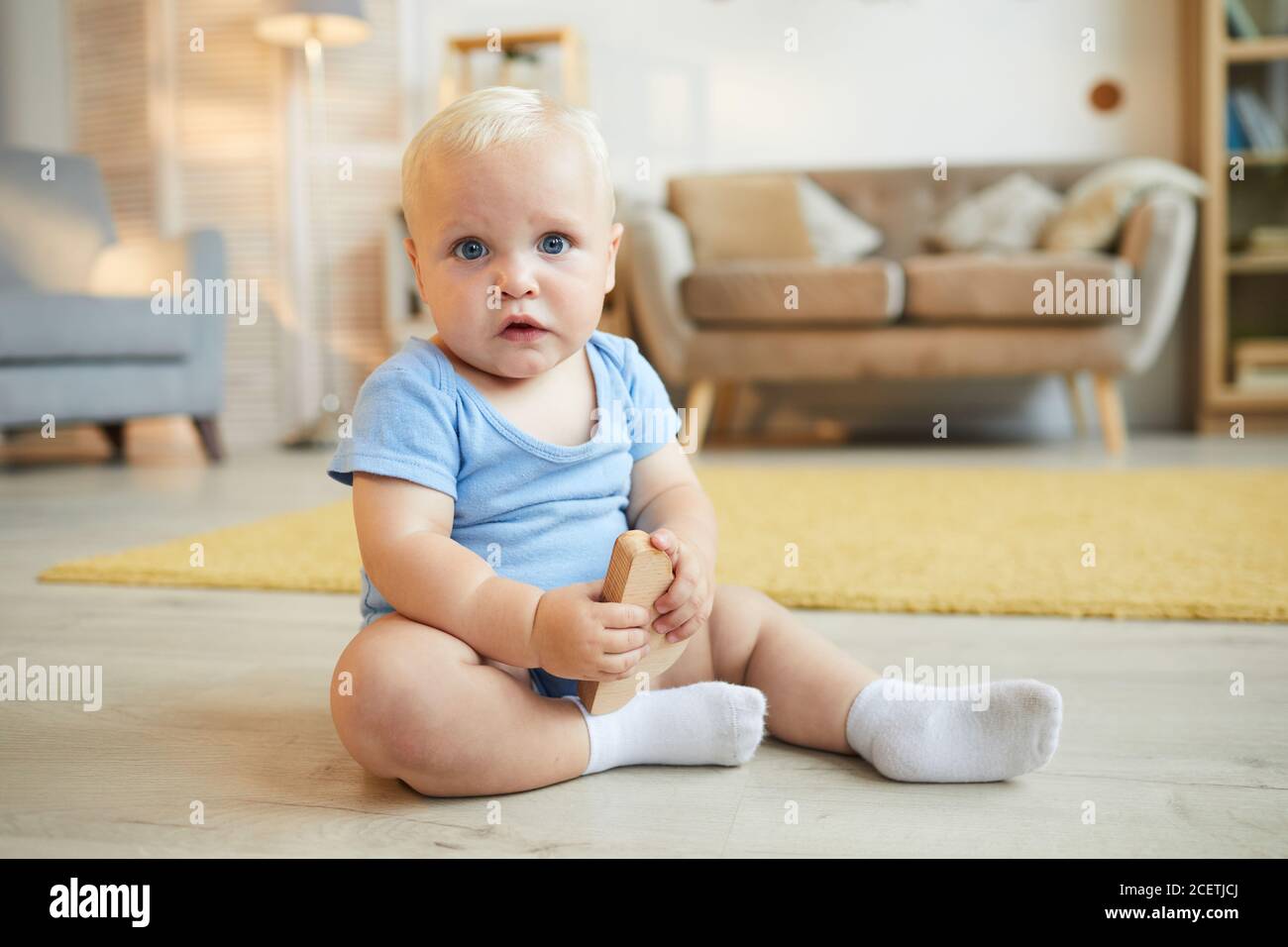Image horizontale du corps de bébé mignon avec des cheveux blonds assis sur le sol dans le salon, regardant l'appareil photo, l'espace de copie Banque D'Images