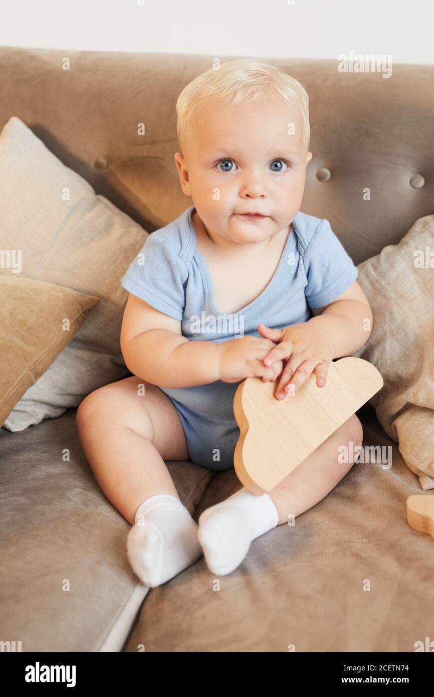 Prise de vue verticale grand angle du corps d'un petit enfant avec cheveux blonds et yeux bleus assis sur le canapé avec bois jouet regardant l'appareil photo Banque D'Images