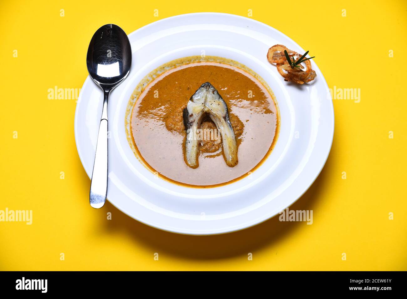 Soupe à la crème ou purée brune sur fond jaune. Soupe de poisson avec un morceau de poisson et des rondelles d'oignon frits sur une assiette blanche. Plat de restaurant avec une cuillère métallique. Concept de cuisine française. Banque D'Images