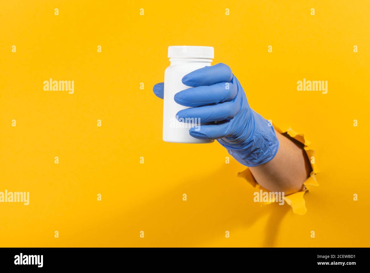 Main dans un gant en latex tenant une bouteille blanche de pilules à travers le mur jaune déchiré Banque D'Images