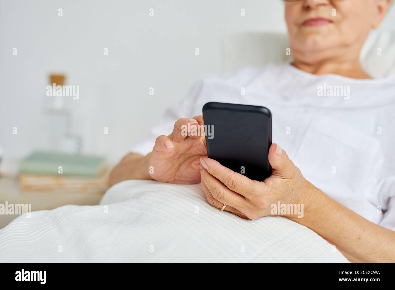 Femme moderne, vêtue de vêtements blancs, se détendant au lit envoyer des sms à l'hôpital à l'aide de son smartphone Banque D'Images