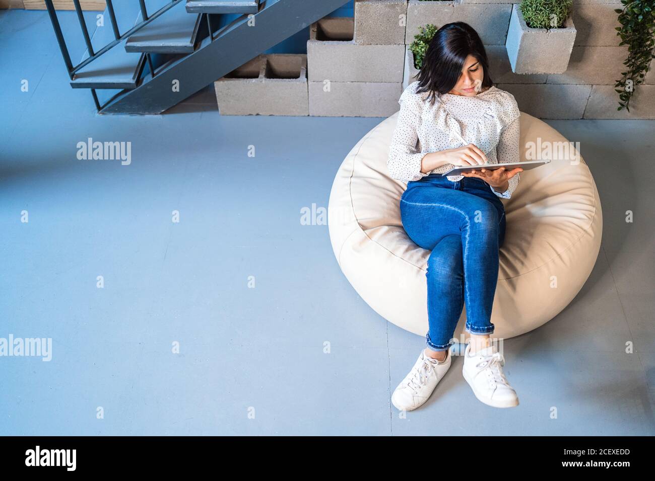 Vue de dessus sur toute la longueur de jeune femme designer focalisée dans vêtements décontractés sur un confortable pouf et avec une tablette graphique tout en travaillant dans un espace de travail créatif contemporain Banque D'Images