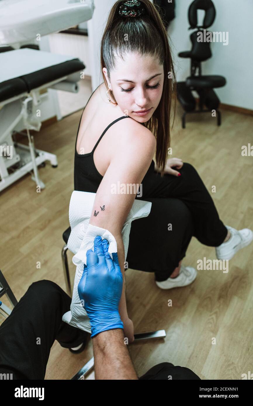 Vue arrière crop anonyme homme tatoueur en latex gants essuyant tatouage fraîchement préparé sur le bras de la cliente avec serviette en salon de coiffure Banque D'Images Vue arrière crop anonyme homme tatoueur en latex gants essuyant tatouage fraîchement préparé sur le bras de la cliente avec serviette en salon de coiffure Banque D'Images