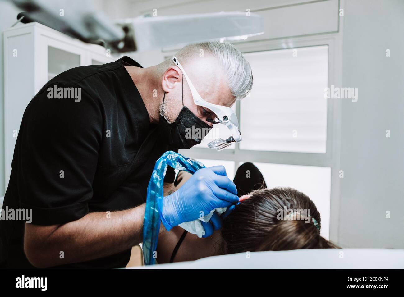 Vue latérale du tatoueur mâle adulte dans le masque de protection et lunettes médicales avec machine de tatouage faisant micro tatouage sur l'oreille de client féminin Banque D'Images