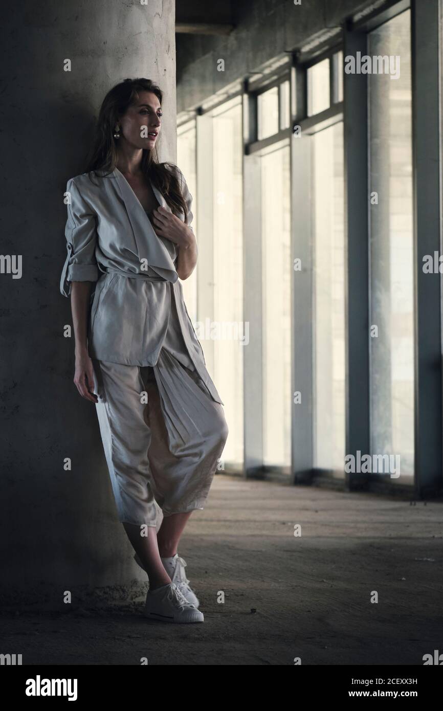 Corps complet de la jeune femme calme dans une élégante veste grise et le pantalon se penche contre une colonne en béton et regarde loin avec soin dans le hall d'un bâtiment contemporain Banque D'Images