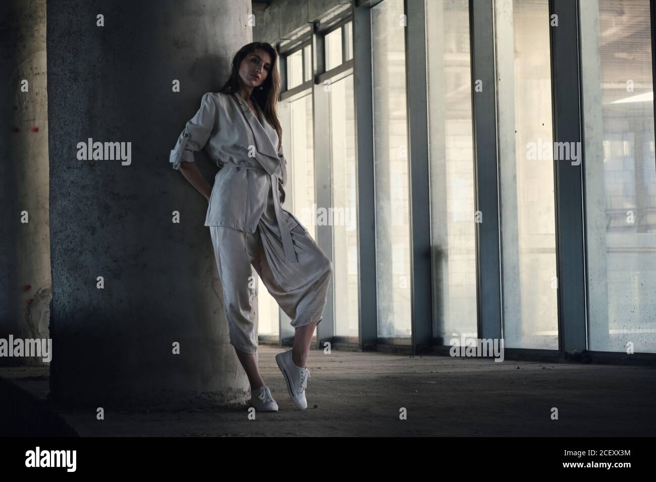 Corps complet de la jeune femme calme dans une élégante veste grise et le pantalon appuyé contre une colonne de béton et regardant l'appareil photo dans le hall d'un bâtiment contemporain Banque D'Images