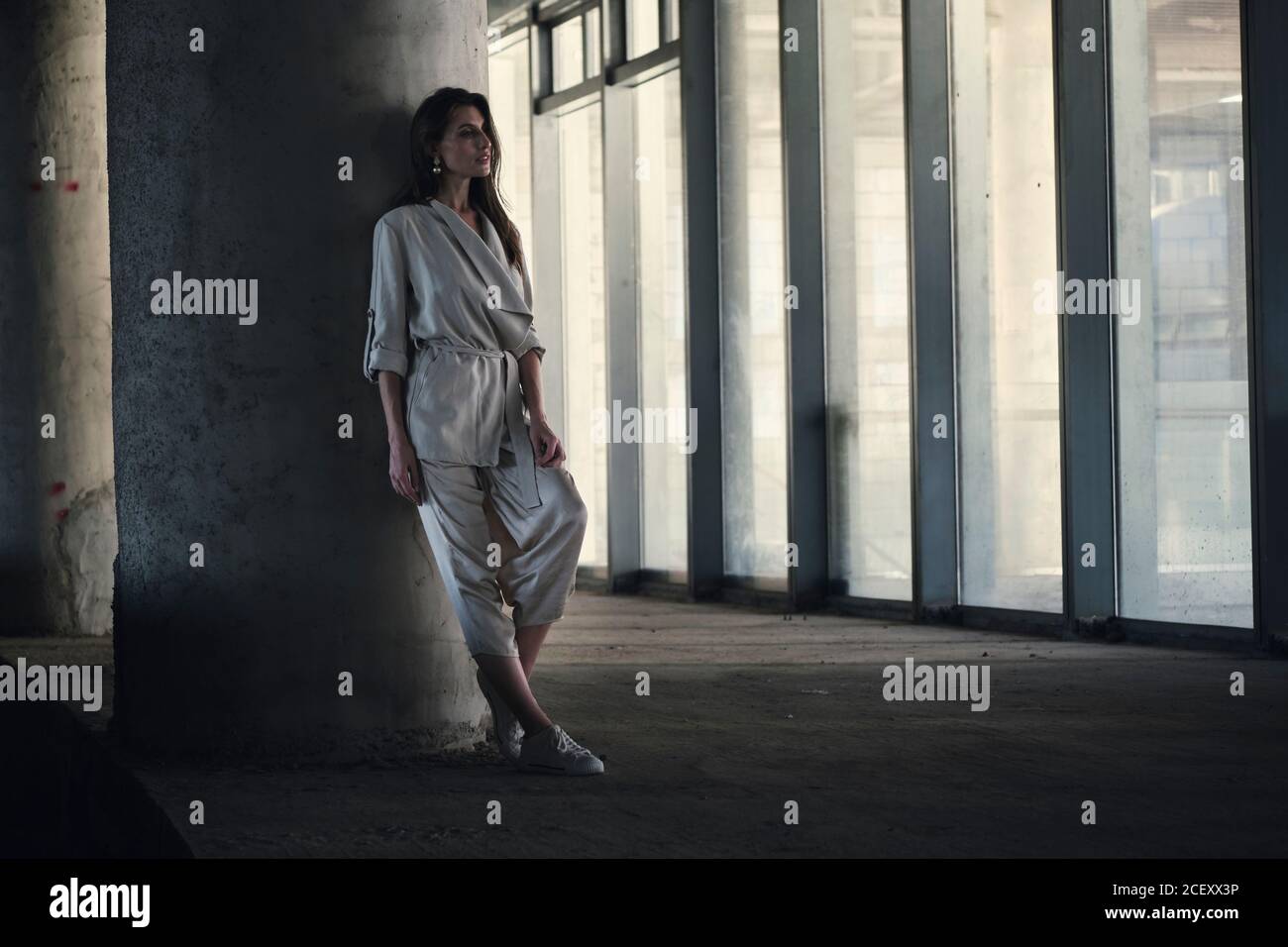 Corps complet de la jeune femme calme dans une élégante veste grise et le pantalon se penche contre une colonne en béton et regarde loin avec soin dans le hall d'un bâtiment contemporain Banque D'Images