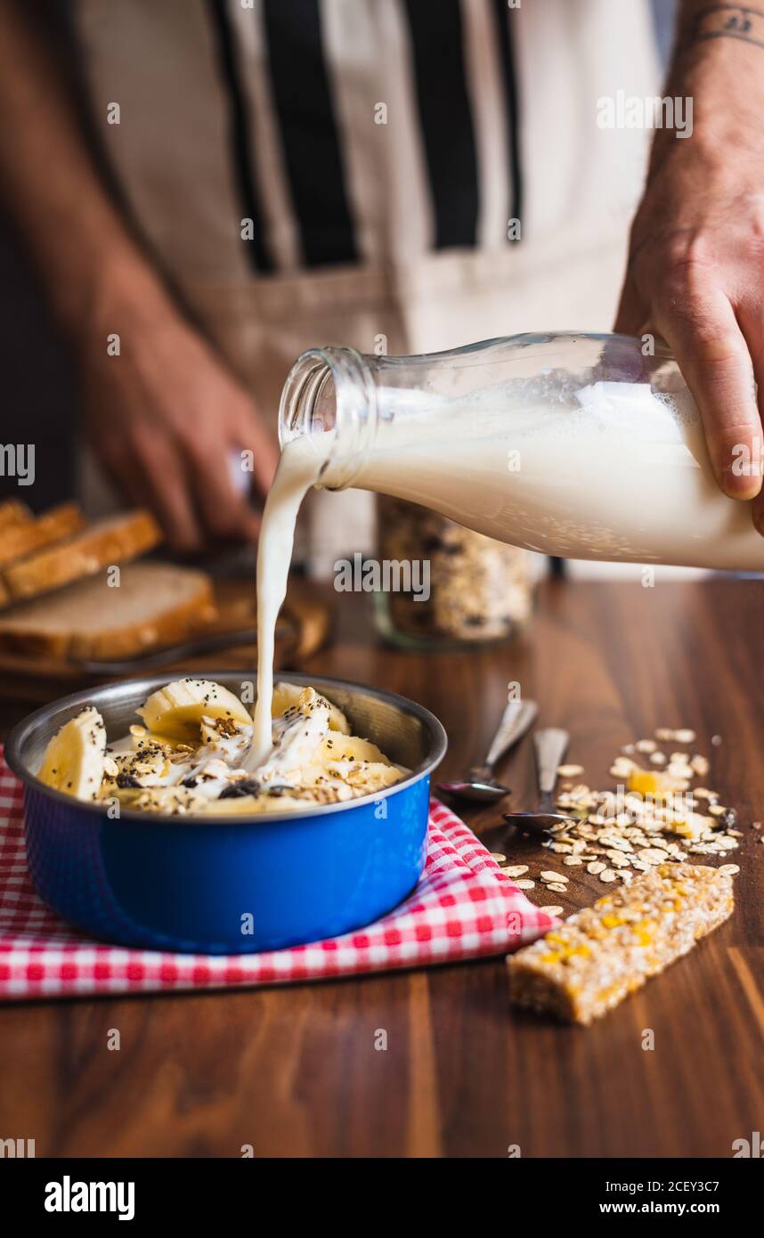 Rognez la femme de ménage anonyme en versant du yogourt de la bouteille de verre dans le bol avec des céréales et de la banane tout en préparant un petit déjeuner sain à la maison cuisine Banque D'Images