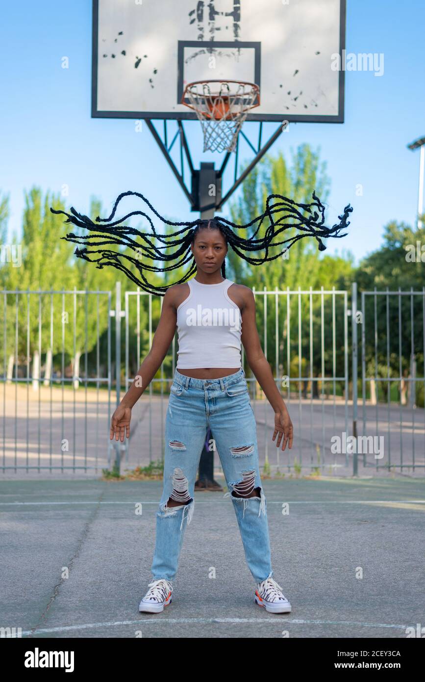 Jeune femme afro-américaine pleine corps en jeans déchirés tendance et le dessus blanc avec des braïdes de tissage debout contre le cerceau de basket-ball dans un parc urbain Banque D'Images