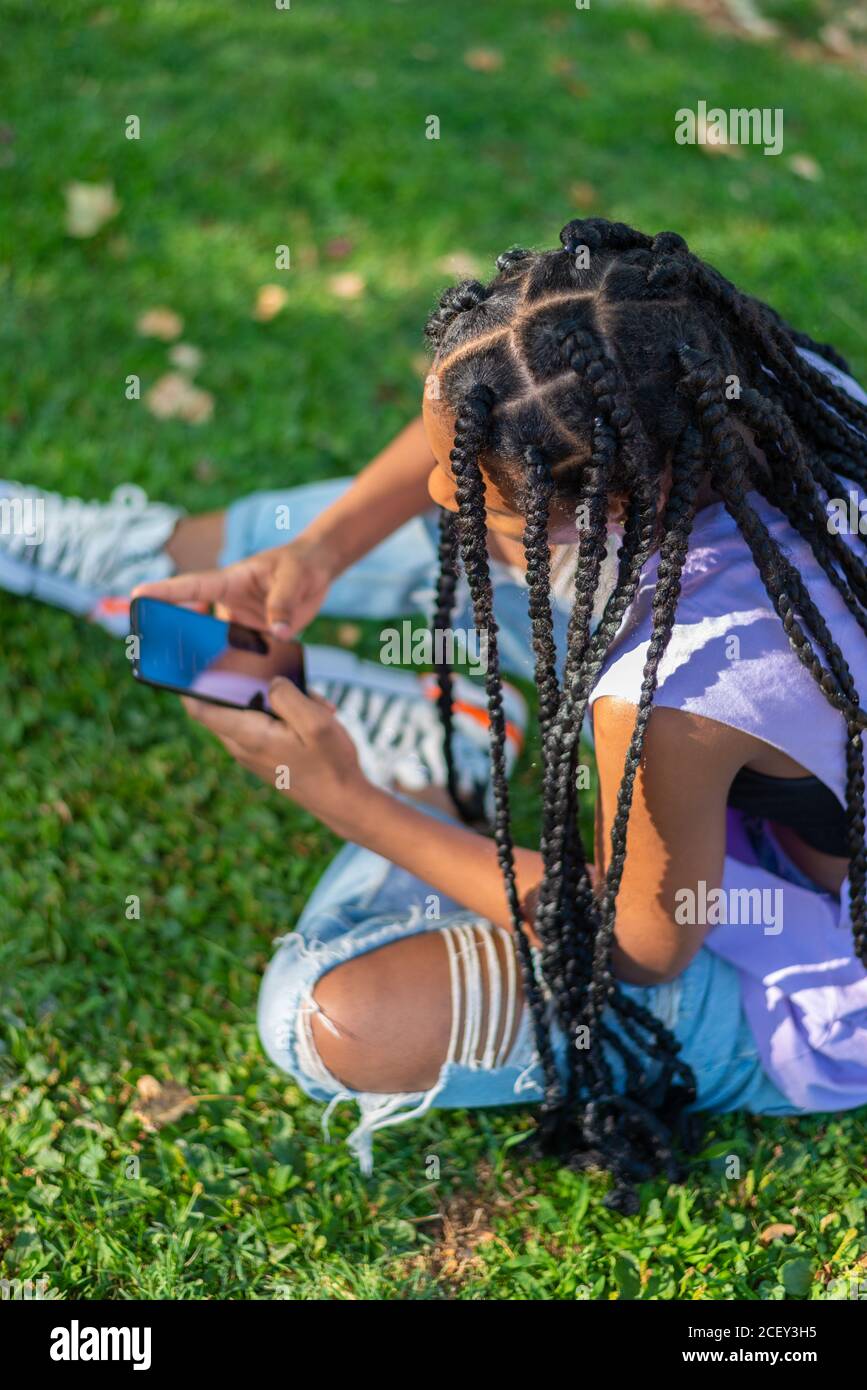 Vue latérale haute angle de l'hippster moderne Afro-américain femme Avec les pigeons afro sur l'herbe verte et le smartphone de navigation pendant que vous vous reposez dans le parc en été Banque D'Images