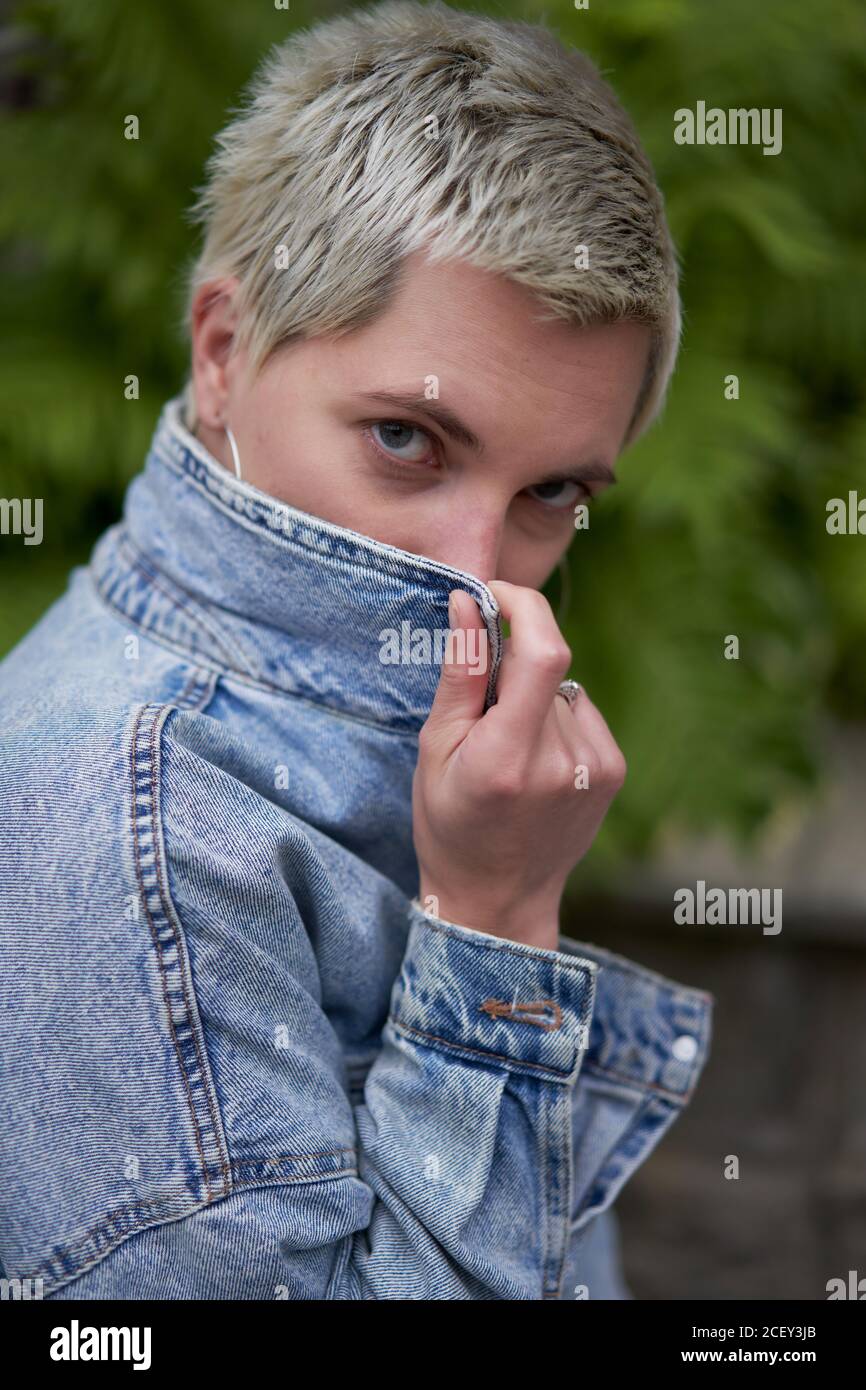 Vue latérale d'une femme confiante dans une tenue tendance street et couvrir le visage avec une veste en denim tout en regardant appareil photo Banque D'Images