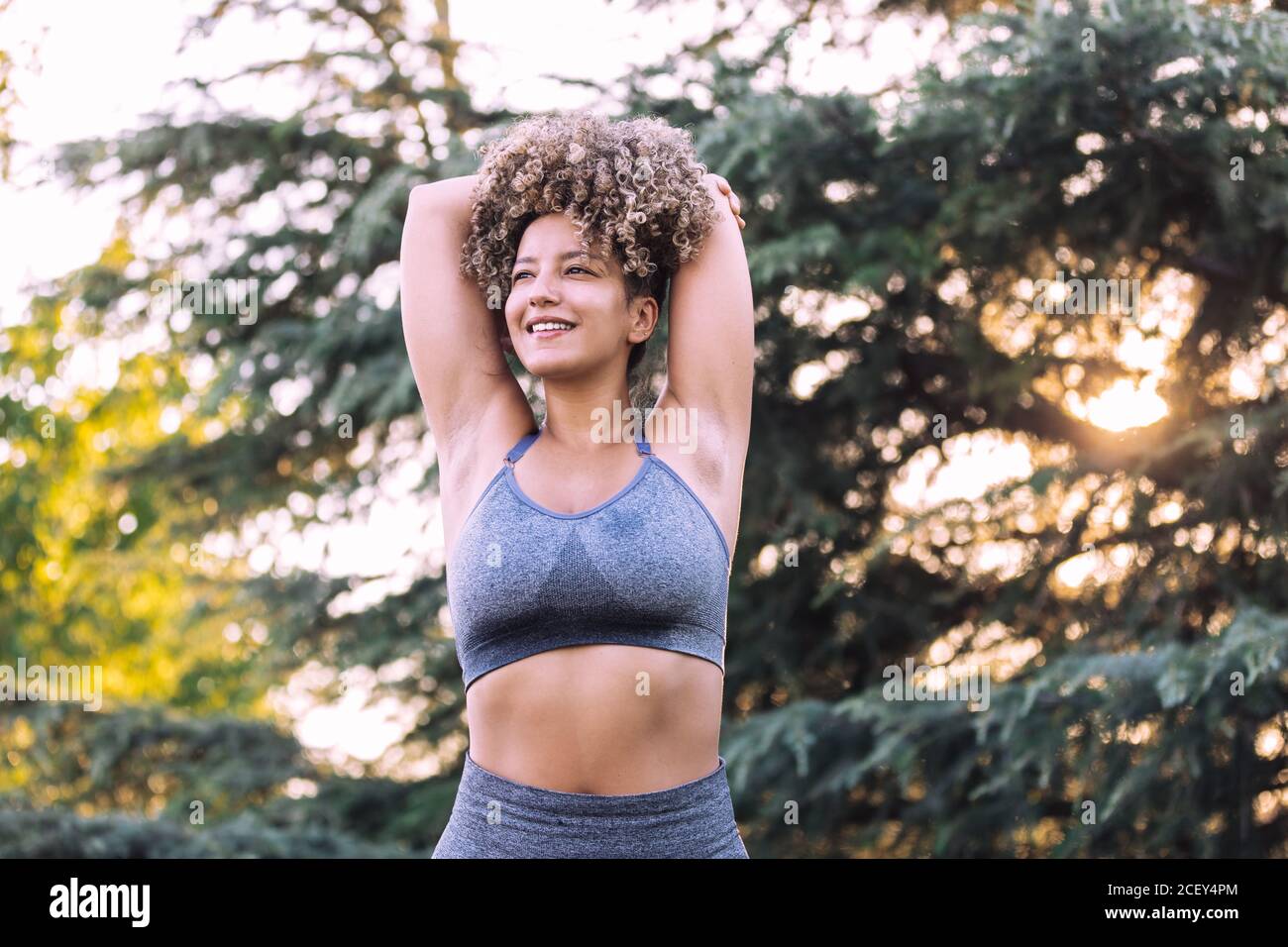 Faible angle de positive énergique jeune femme ethnique avec afro coiffez-vous en portant un haut gris et des leggings qui s'étirent avec les bras derrière vous dirigez-vous à proximité d'arbres verts pendant l'entraînement dans le parc Banque D'Images