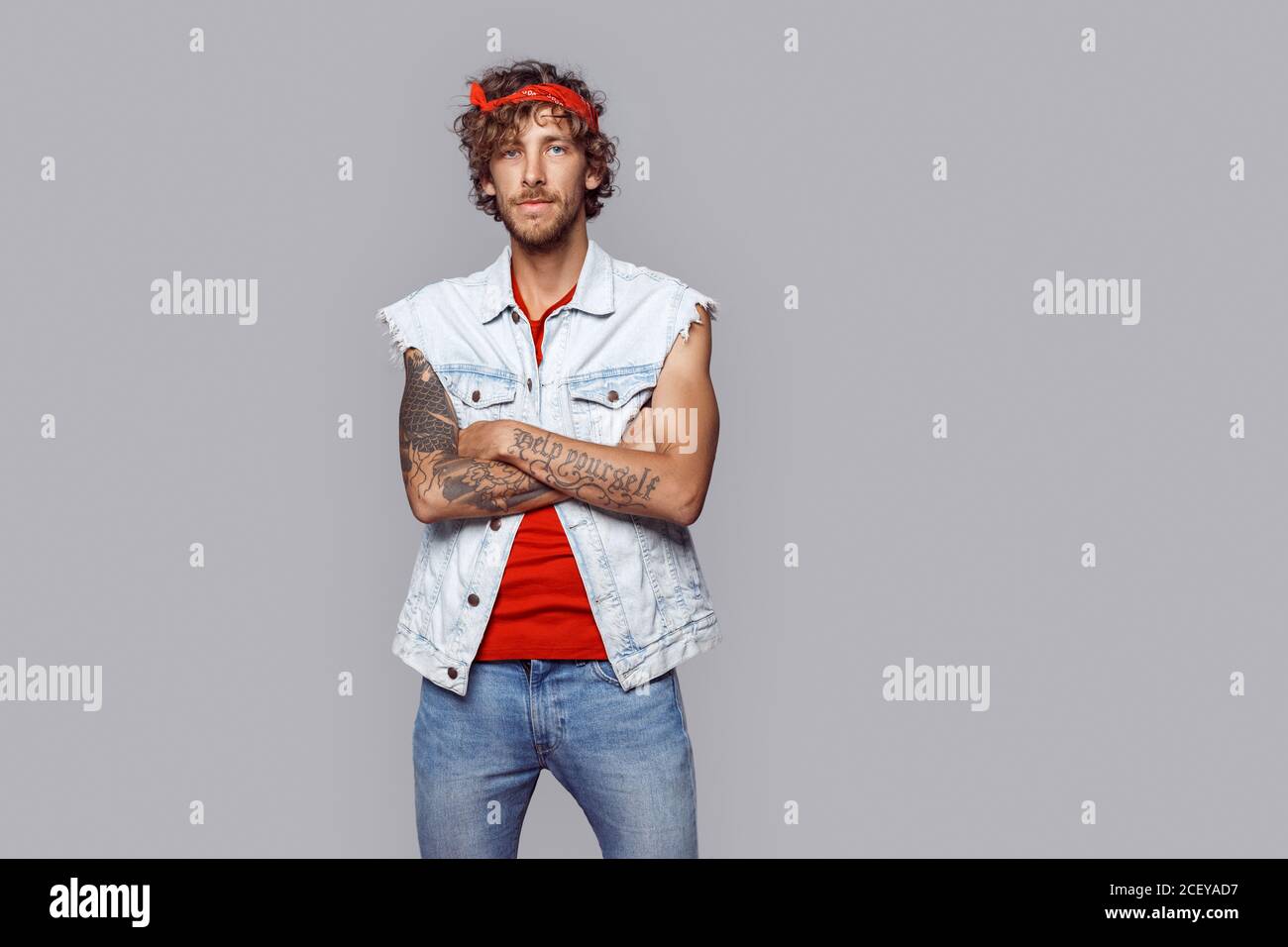 Style libre. Homme aux cheveux bouclés dans jean veste sans manches et  bandeau debout isolé sur les bras gris croisés sourire détendu Photo Stock  - Alamy