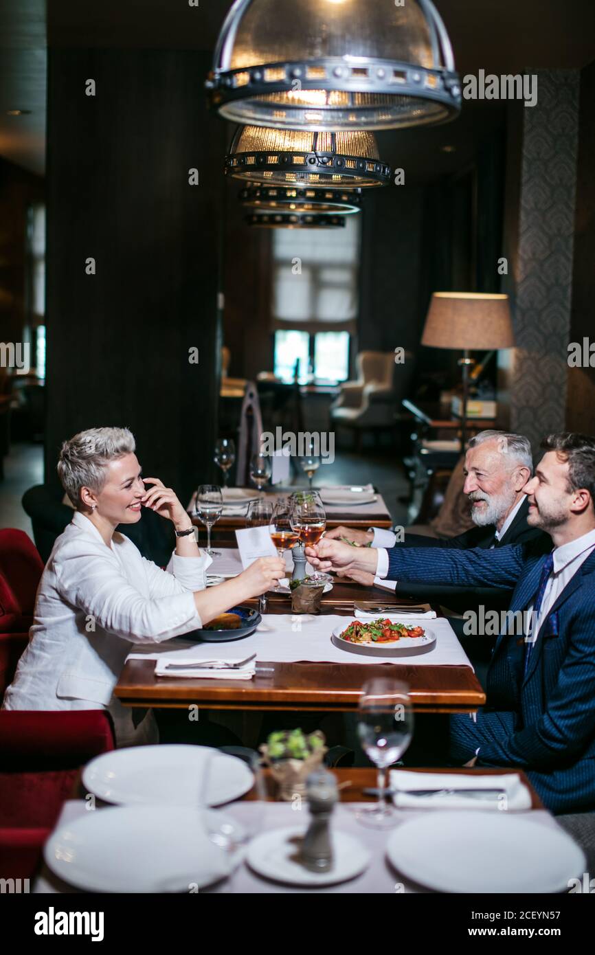 Des partenaires d'affaires joyeux et satisfaits dans des vêtements habillés, asseyez-vous à table, levez des verres à vin avec des visages joyeux, célébrez avec passion l'anniversaire Banque D'Images