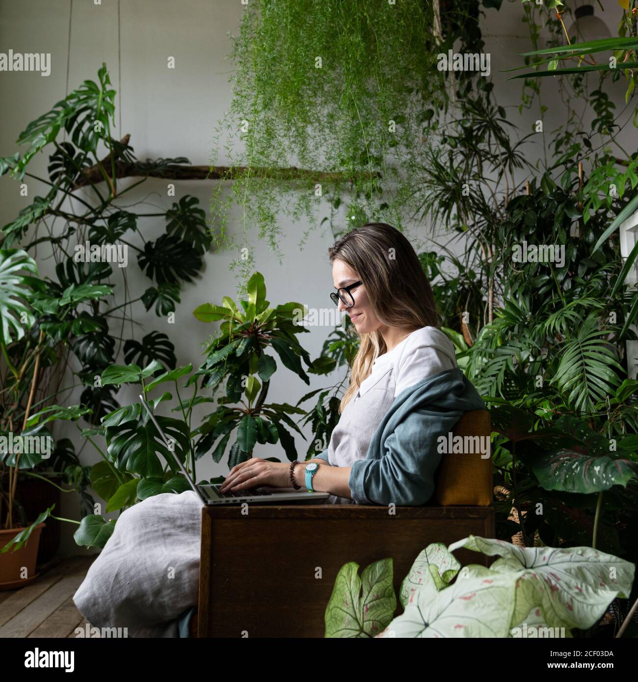 Gros plan portrait d'une femme souriante gardenier en lunettes porter une robe de lin, s'asseoir sur une chaise en serre, utiliser un ordinateur portable et parler sur Internet Banque D'Images