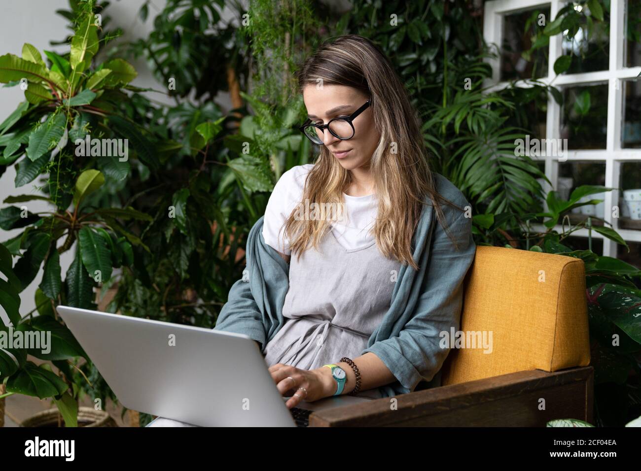 Femme jardinier portant une robe de lin, assise sur une chaise dans une maison verte, travaillant sur un ordinateur portable entouré de plantes. Jardin à la maison, travail à distance, travail à distance. Banque D'Images