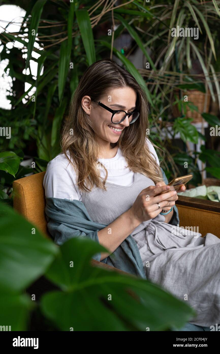Gros plan de sourire positif femme jardinier porter la robe de lin, assis sur une chaise dans la maison verte, se reposer, en utilisant le smartphone et l'envoi de message Banque D'Images