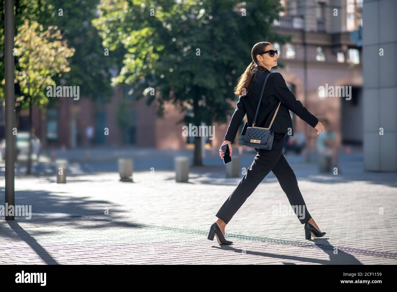 Élégante femme aux cheveux foncés dans des lunettes de soleil traversant la rue Banque D'Images