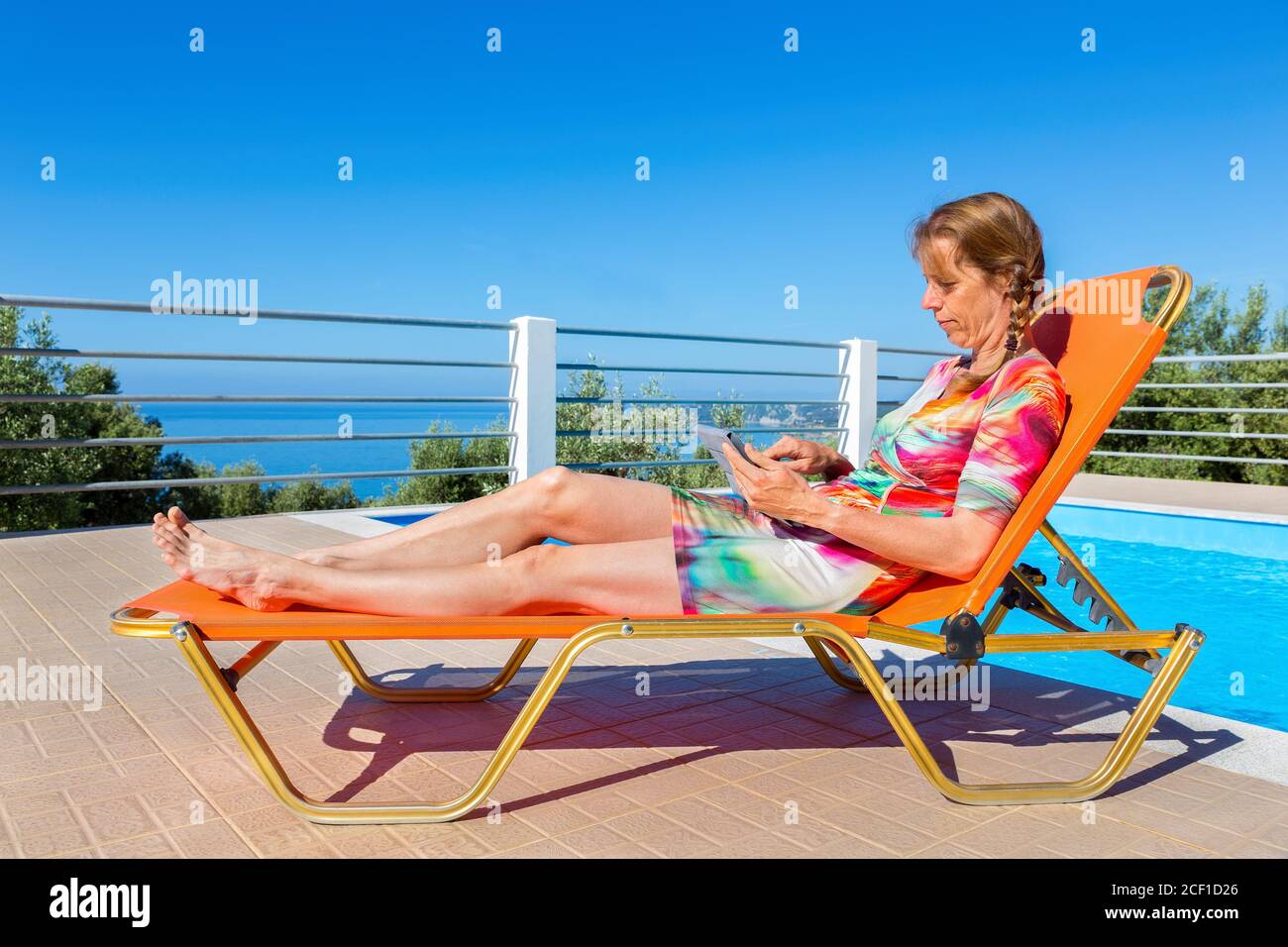 Femme hollandaise d'âge moyen allongé sur une chaise longue orange pour lire un comprimé près de la piscine Banque D'Images