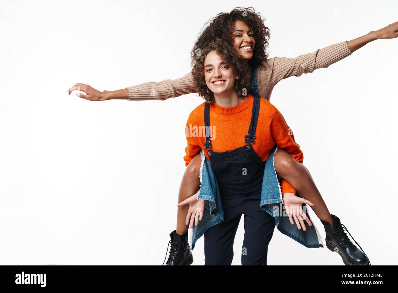 Image de l'incroyable optimiste heureux multiracial filles amis en denim combinaisons isolées sur fond de mur blanc ayant le plaisir. Banque D'Images