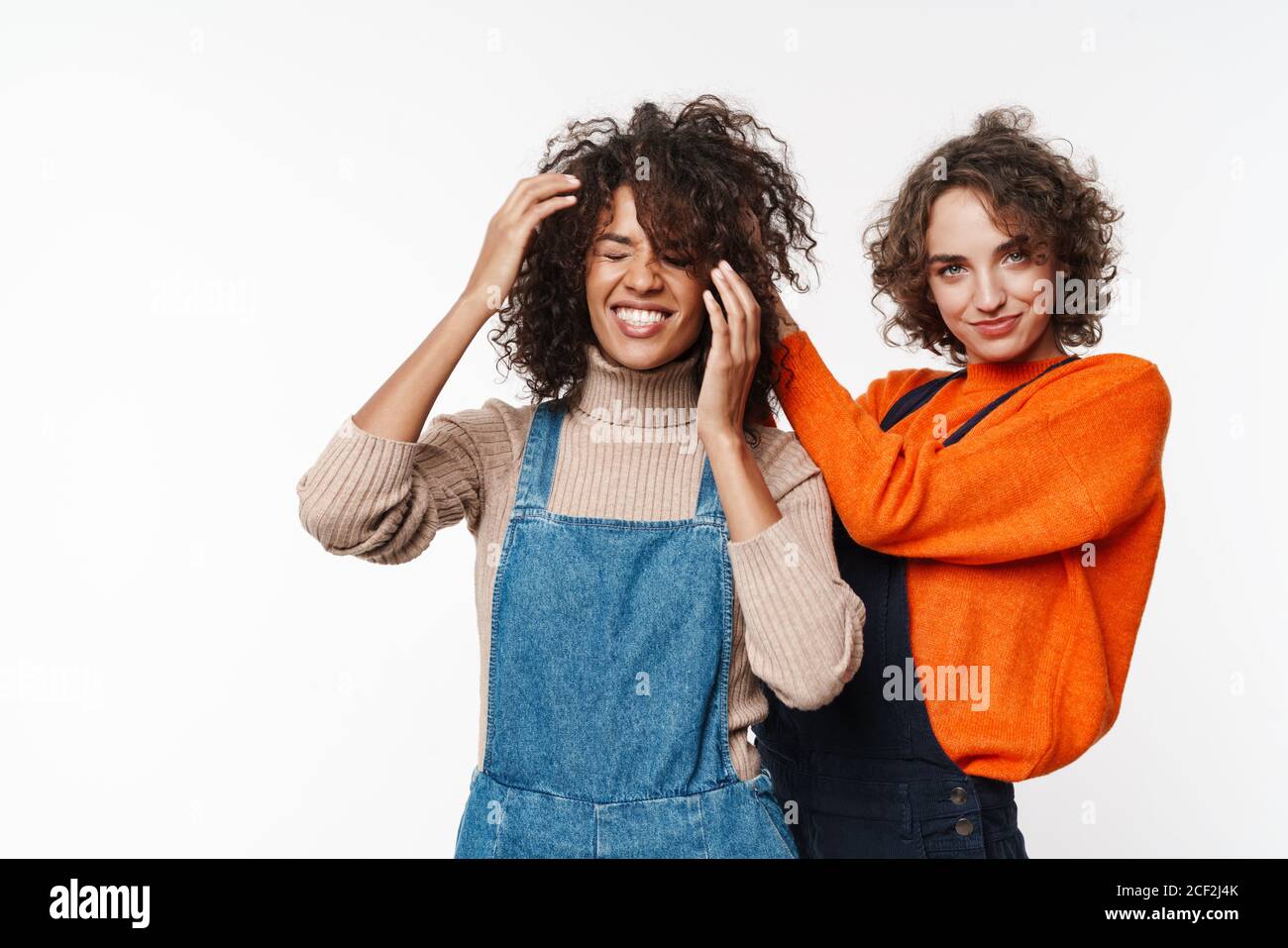 Image d'une belle gaie heureuse et optimiste deux filles multiraciales amis en jean combinaisons isolées sur fond de mur blanc ayant plaisir de secouer h Banque D'Images