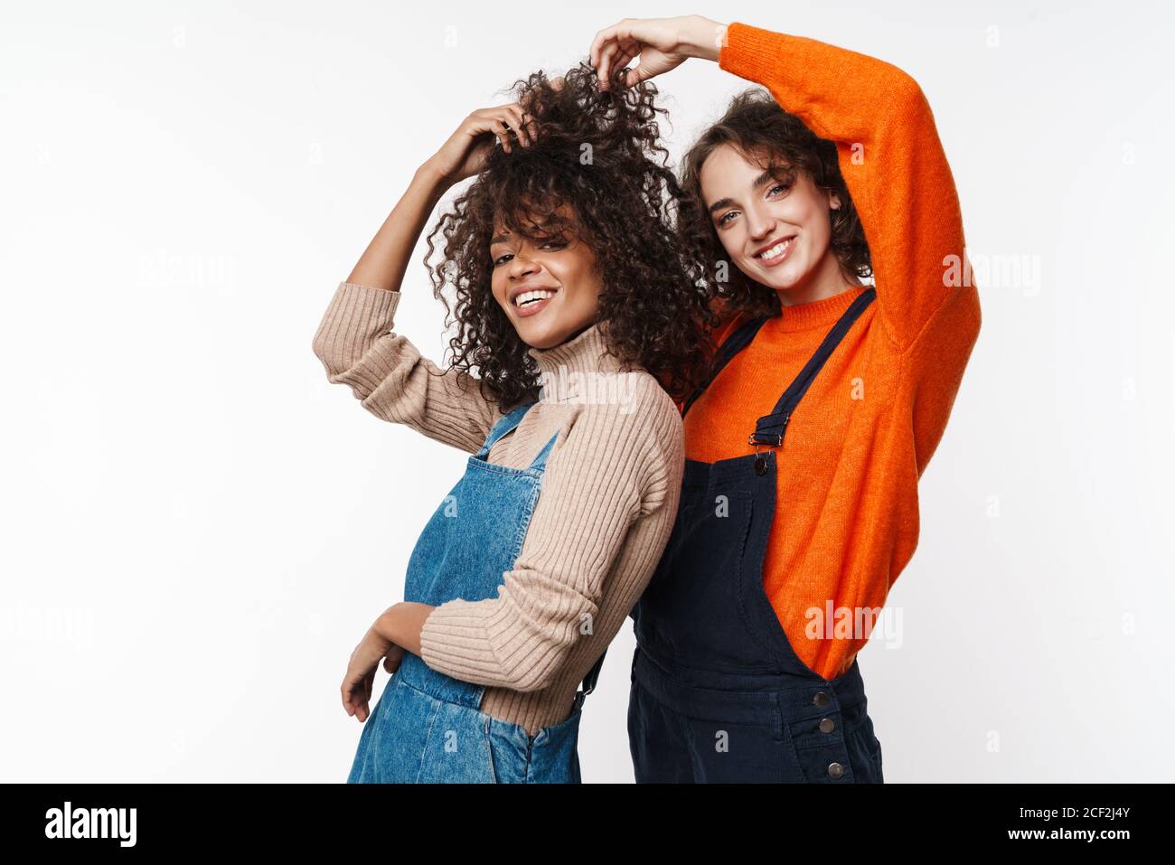 Image d'un joli sourire souriant deux amies multiraciales en combinaisons denim isolées sur fond de mur blanc. Banque D'Images