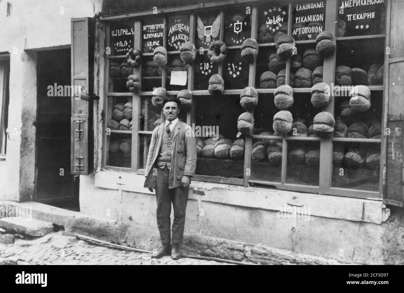 Baker debout devant la boulangerie américaine qui affiche des panneaux en arménien, Ladino (en caractères hébreux), anglais, turc ottoman, grec et russe avec des échantillons de pain attaché aux meneaux, Ortaköy, Istanbul, Turquie, 1922 Banque D'Images