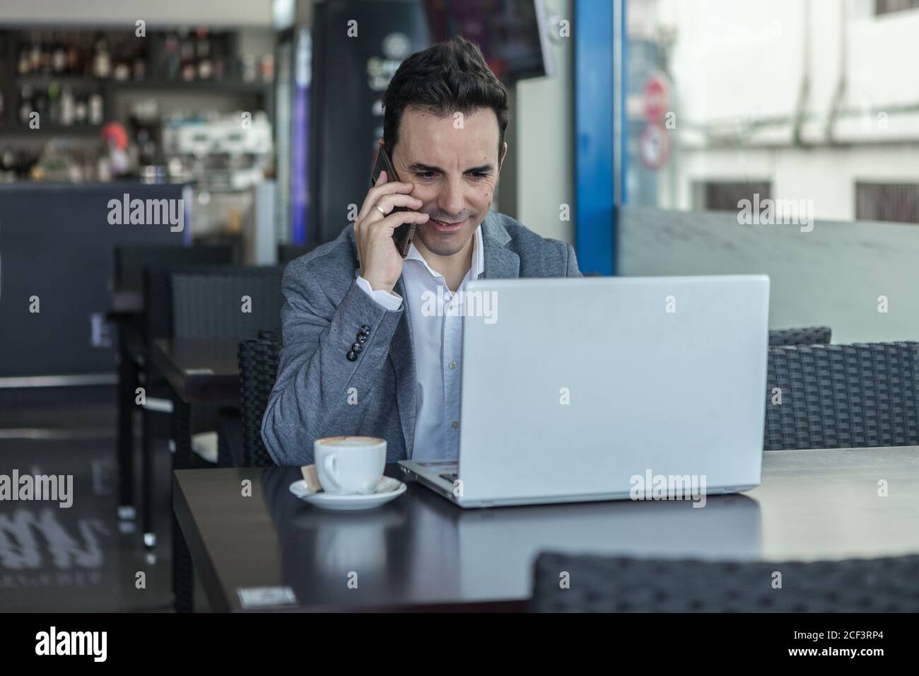 Manager homme adulte souriant dans des vêtements formels regardant l'ordinateur portable écran et conversation sur smartphone tout en travaillant en ligne dans les modernes café Banque D'Images