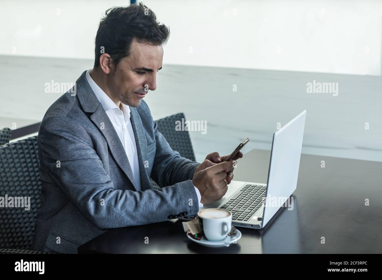 Vue latérale d'un homme d'affaires adulte sérieux en costume habillé assis à la table avec une tasse de café et un ordinateur portable et vérification les informations sur le téléphone portable pendant que le Banque D'Images