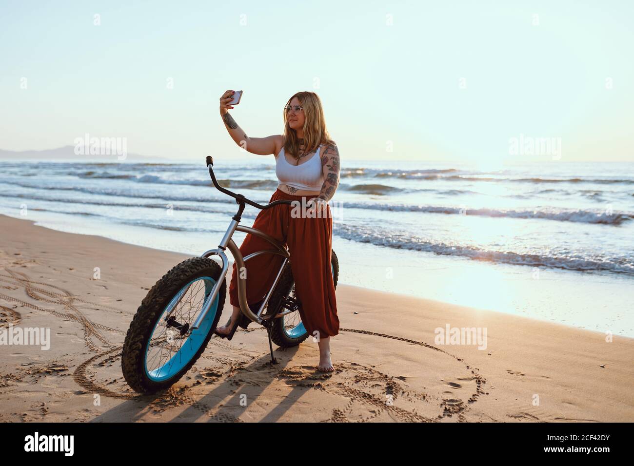 Joyeuse femme en train de prendre un selfie sur un smartphone tout en se reposant à vélo sur une rive ensoleillée Banque D'Images