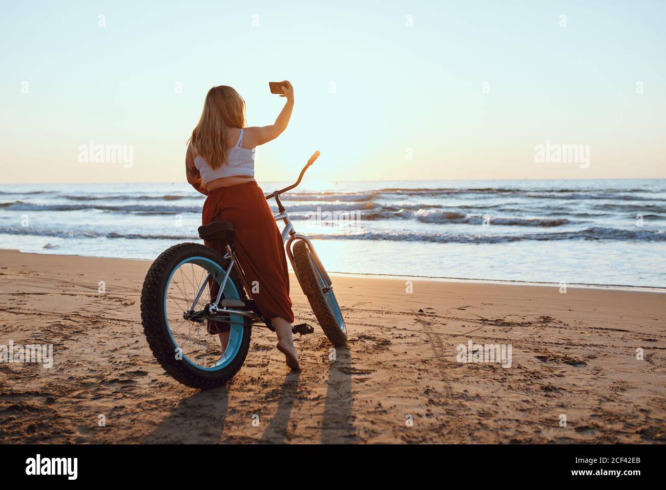 Joyeuse femme en train de prendre un selfie sur un smartphone tout en se reposant à vélo sur une rive ensoleillée Banque D'Images