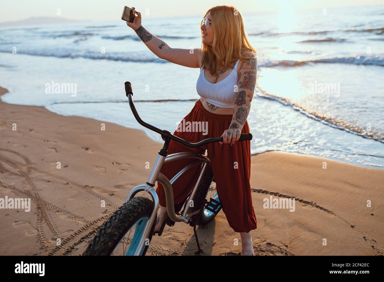 Joyeuse femme en train de prendre un selfie sur un smartphone tout en se reposant à vélo sur une rive ensoleillée Banque D'Images
