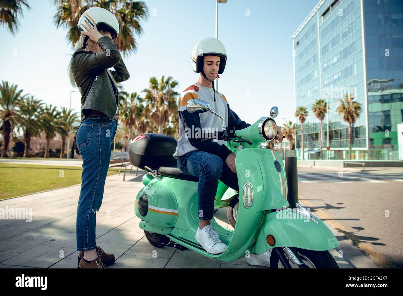 Vue latérale d'un beau homme dans un casque assis et débutant un scooter vert et d'une jeune femme dans une veste noire portant un casque Banque D'Images