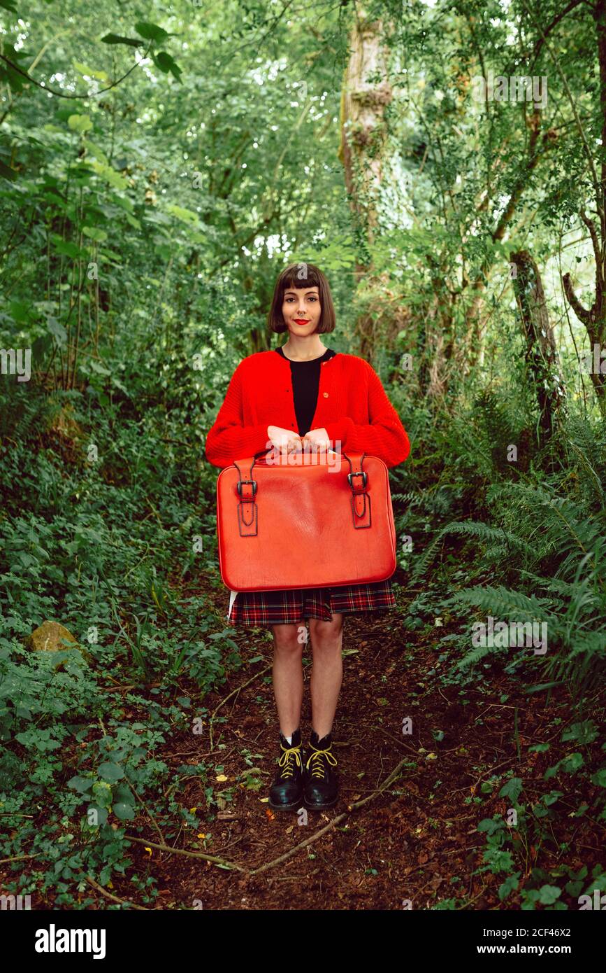 Jeune femme élégante en chandail rouge avec grande valise rouge debout regardant directement l'appareil photo dans une forêt dense Banque D'Images