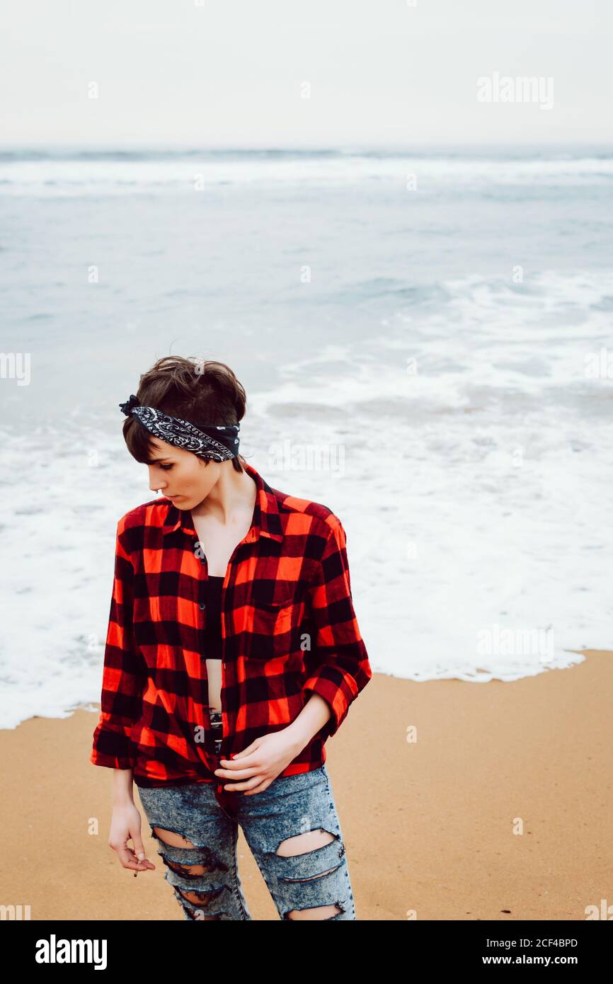 De dessus de la femme pensive malheureuse dans la chemise à carreaux et jean déchiré debout avec une cigarette à la main sur une plage de sable avec la mer de tempête en arrière-plan Banque D'Images