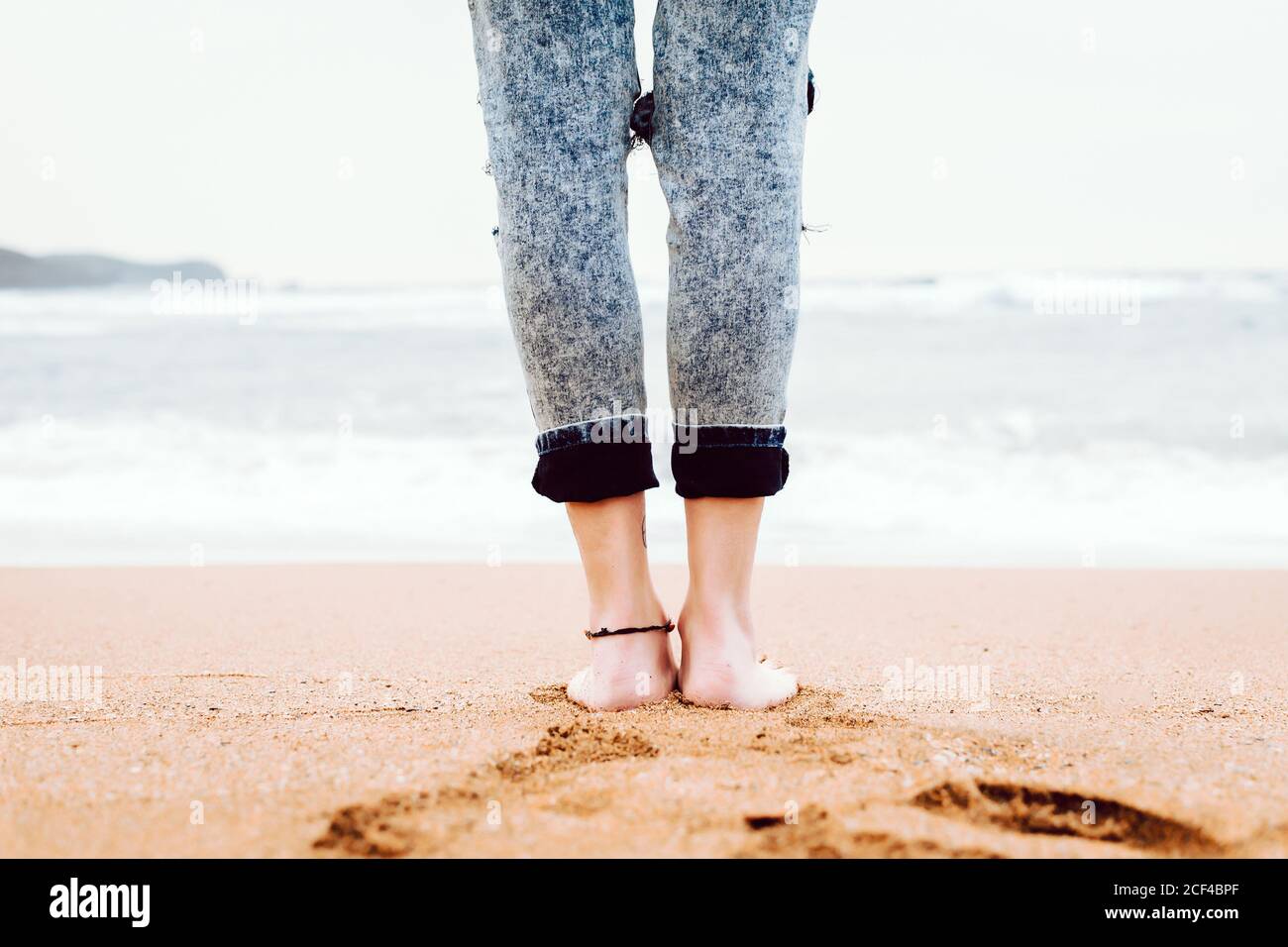 Vue arrière des jambes de la femme pieds nus portant du denim sur une plage de sable avec une mer orageux et un ciel gris en arrière-plan Banque D'Images