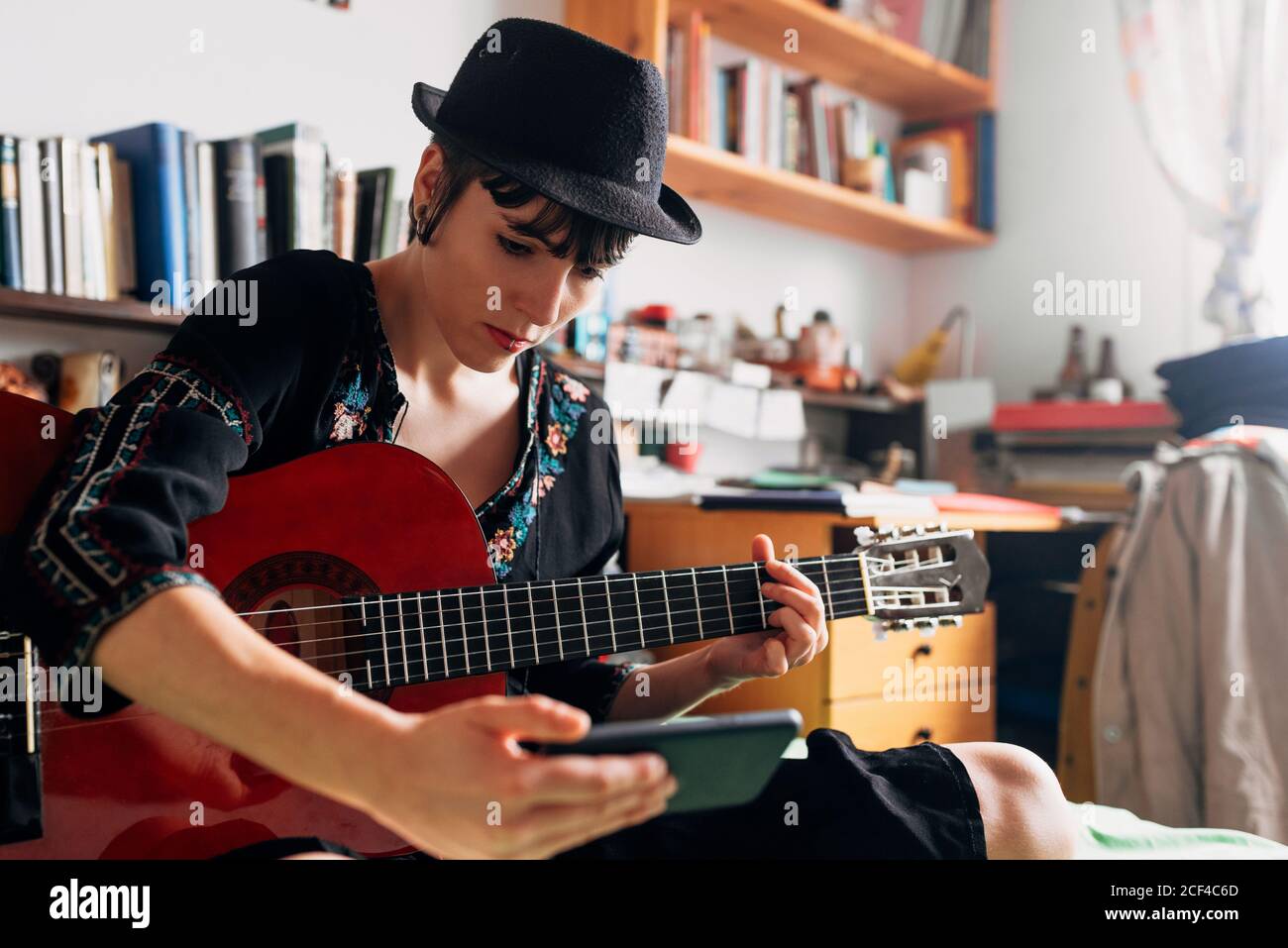 Femme en tenue tendance et chapeau assis sur le lit avec guitare acoustique et lecture de messages sur le téléphone portable tout en refroidissant pendant week-end Banque D'Images