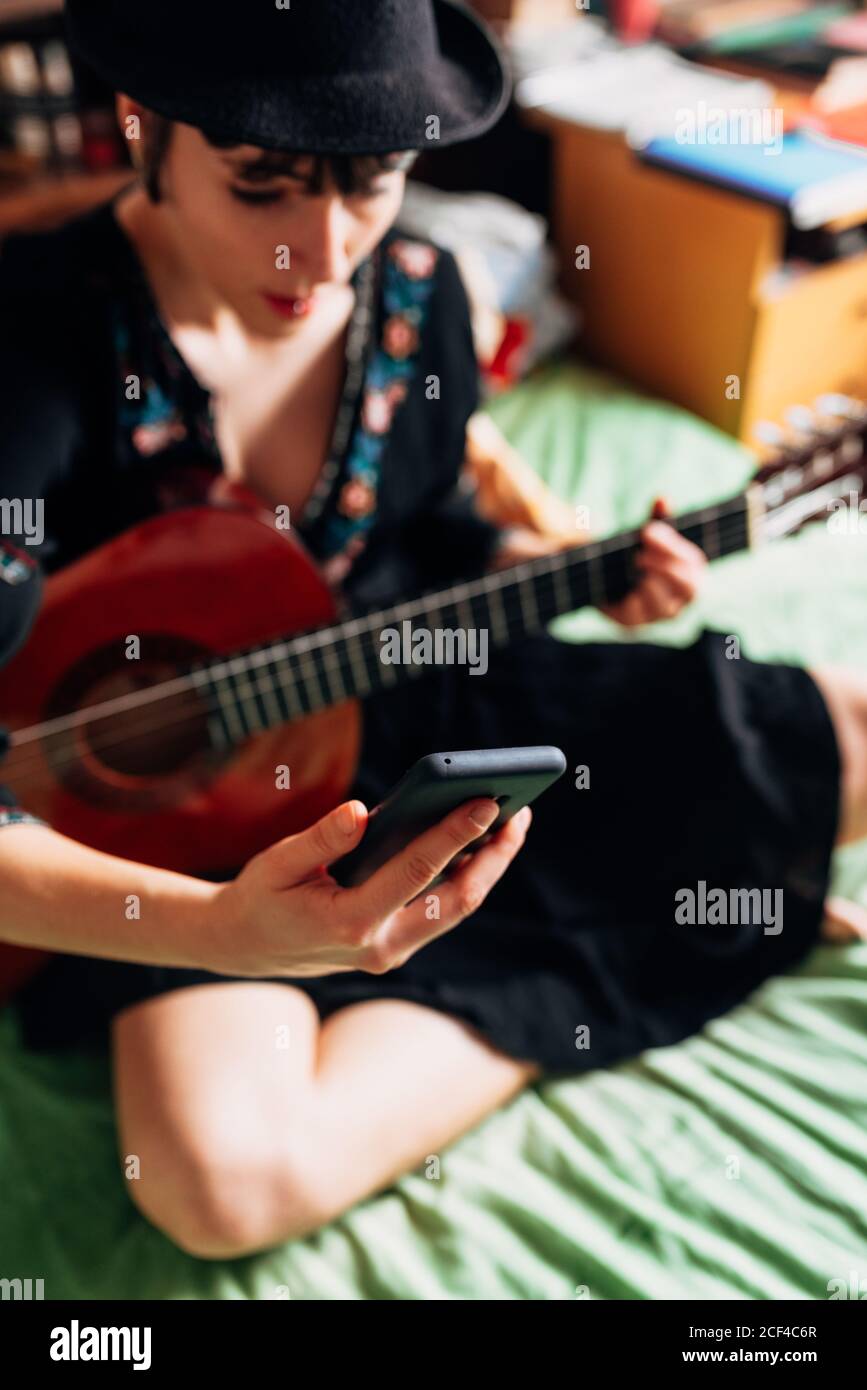 Femme en tenue tendance et chapeau assis sur le lit avec guitare acoustique et lecture de messages sur le téléphone portable tout en refroidissant pendant week-end Banque D'Images
