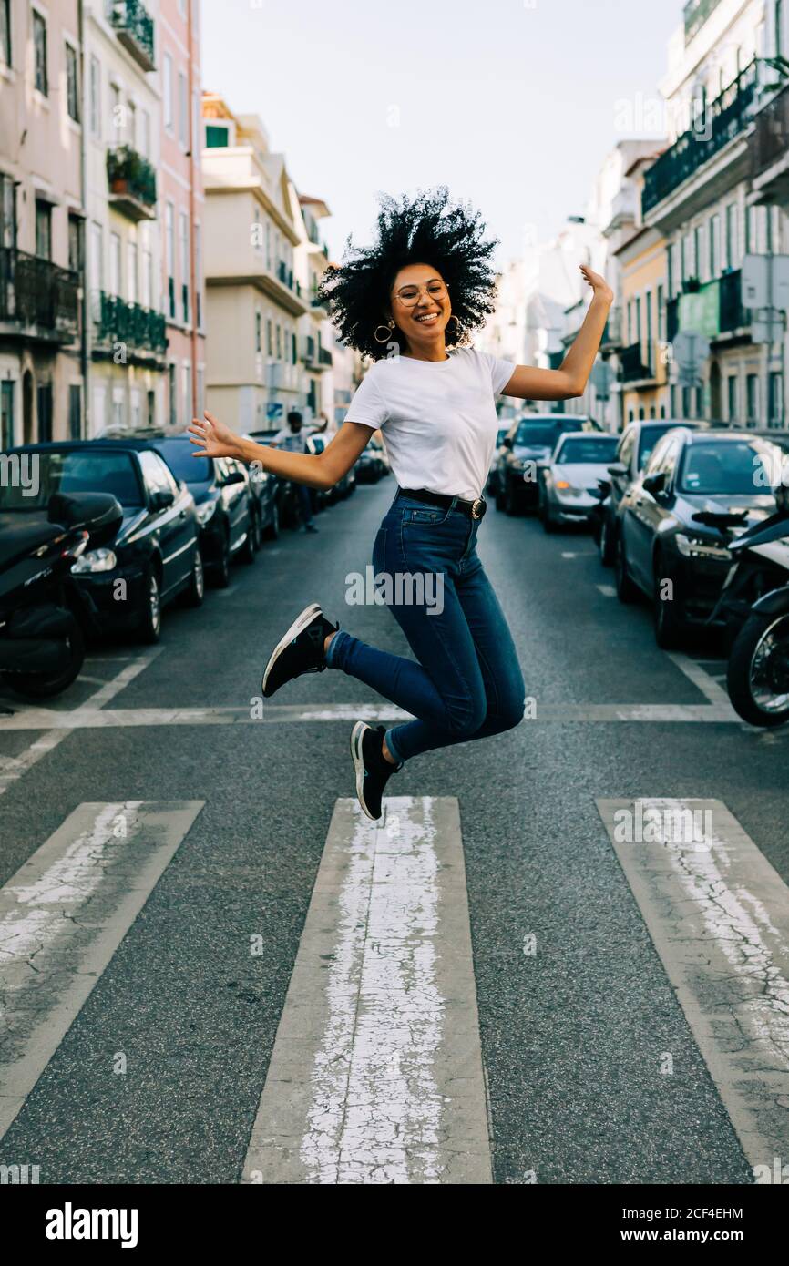 Jeune femme afro-américaine joyeuse en jeans et t-shirt blanc sauter pour la joie dans la rue de la ville le jour Banque D'Images