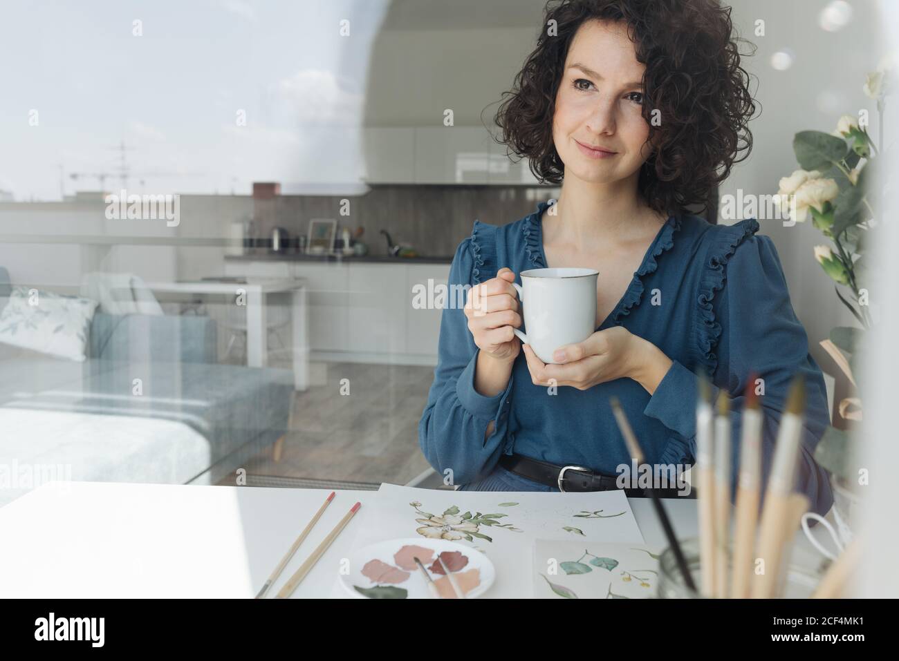 Femme brune pensive artiste assis à table avec une tasse de café et regardant loin dans le lieu de travail Banque D'Images
